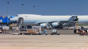 Boeing 747 freighter with its nose cargo door open on a sunny airport apron, cargo pallets and ground crew in front