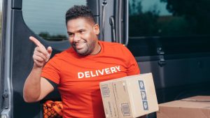 Smiling courier in a red Delivery T-shirt holding a cardboard parcel beside an open delivery van.