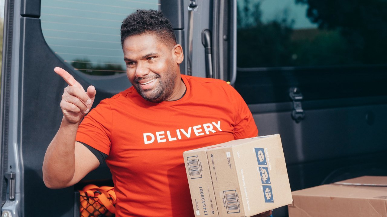 Smiling courier in a red Delivery T-shirt holding a cardboard parcel beside an open delivery van.