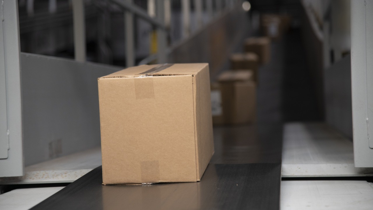 Plain cardboard parcel riding a dark conveyor belt in a parcel sorting hub, with more boxes queued behind it