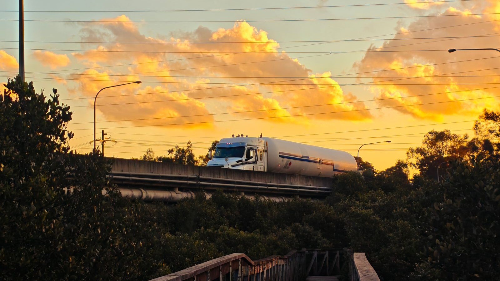 White fuel tanker truck crossing a highway bridge at sunset, with trees and power lines in the foreground