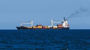 Container ship at sea with exhaust smoke rising from its funnel against a clear blue sky