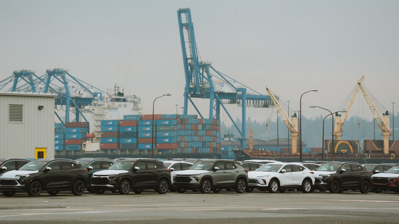 Rows of new SUVs parked at a port vehicle terminal with blue container cranes, stacked containers and a ship behind