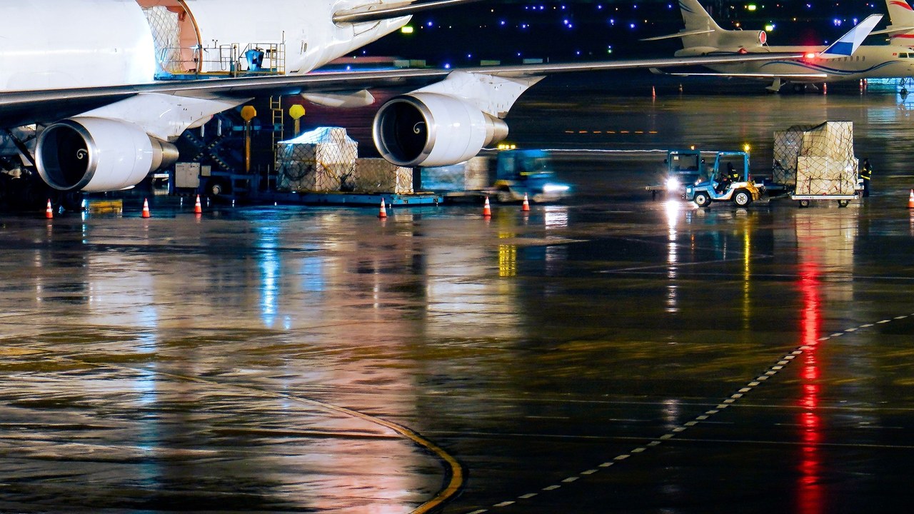Cargo pallets and a tug beside a freighter aircraft on a wet airport apron at night