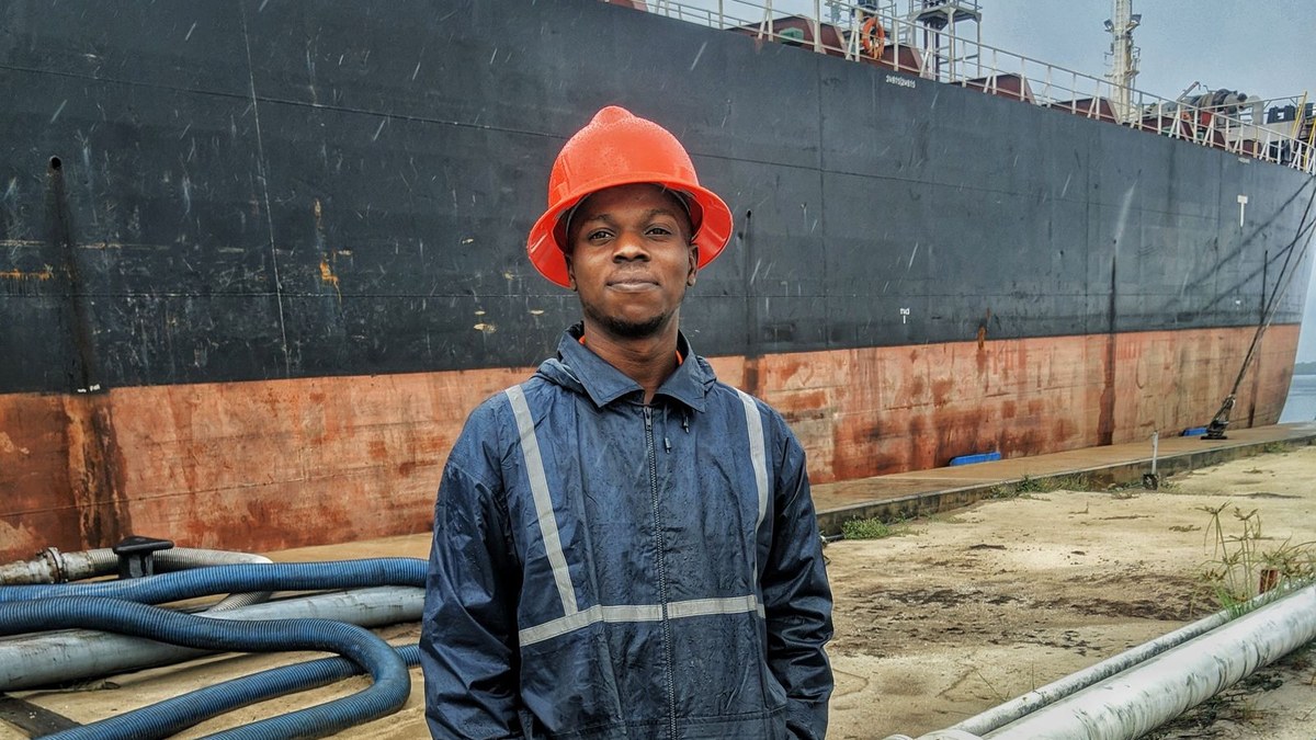 Young dock worker in an orange hard hat and reflective rain gear standing on the quay in front of a large cargo ship