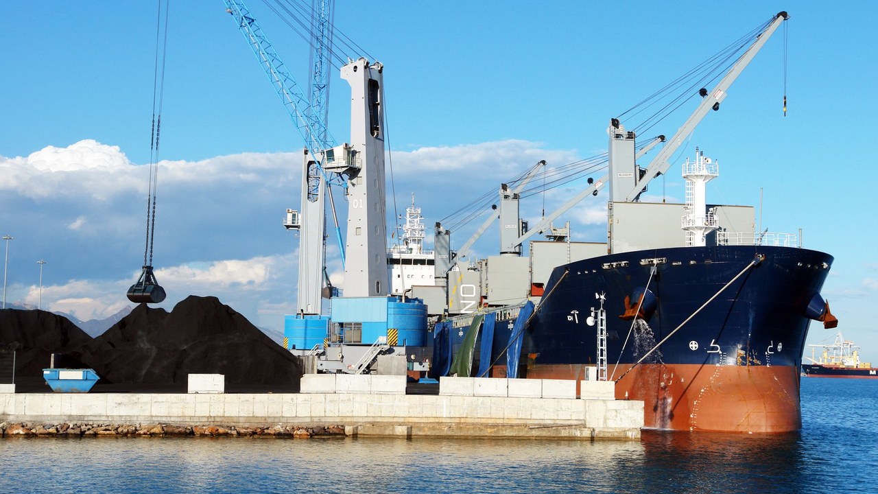 Bulk carrier moored at a port quay while a harbour crane grab loads dark bulk cargo from heaps on the quayside.