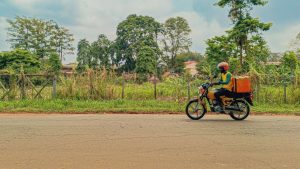 Dispatch rider on a yellow motorcycle with an orange delivery box riding along a tree-lined road in Lagos