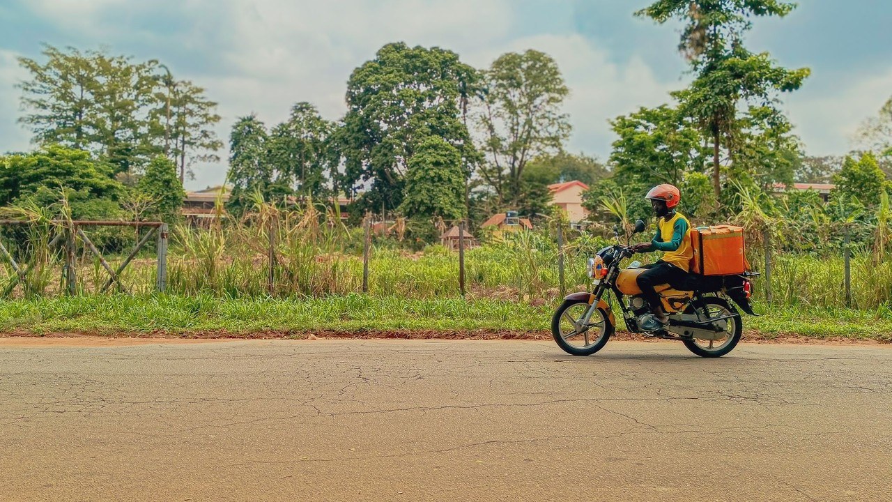 Dispatch rider on a yellow motorcycle with an orange delivery box riding along a tree-lined road in Lagos