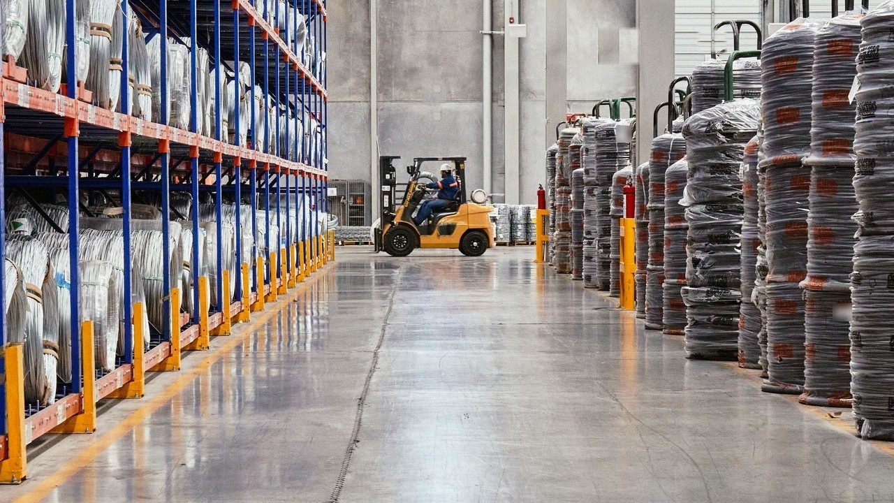 Forklift operator driving down the aisle of a factory raw-materials warehouse between racks of wire coils and tall shrink-wrapped stacks
