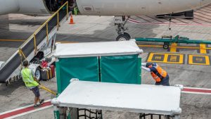 Ground crew pushing covered baggage and cargo carts towards a belt loader at the hold of a passenger aircraft