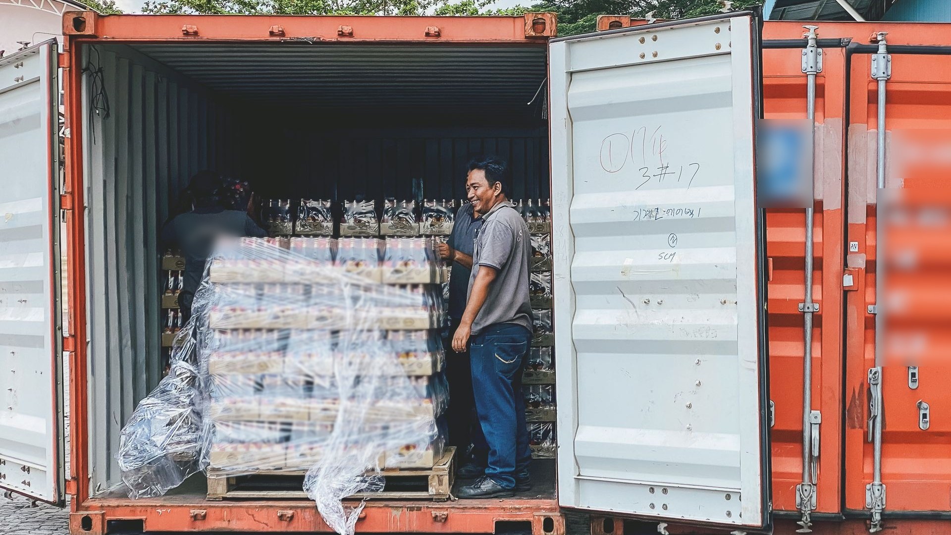 Workers loading a shrink-wrapped pallet of bottled food products into an orange shipping container for export