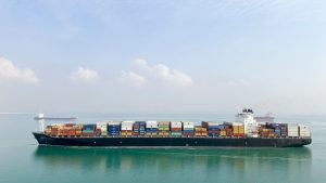 Fully loaded container ship sailing across calm turquoise sea under a hazy sky